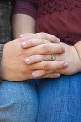 Young couple holding hands with entwined fingers on their engagement day. Polished nails on the female hand. Both are wearing knees.  Hands are resting on the knees. 