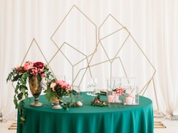 Banquet table decor. On an emerald green tablecloth there is a copper vase with pink and red peonies, flowers in metal geometric stands, candles in glass boxes. In the background is a golden geometric