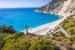 Happy woman standing on top of a rock, raising hands with an exciting feeling of freedom, looking at Myrtos Beach. Cephalonia island, Greece