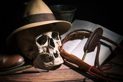 Adventure and archeology concept. Skull with fedora hat, bullwhip, book, quill, shoe, mortyr and magnifying glass on wooden table and black background. Still life