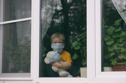 Stay at home quarantine coronavirus pandemic prevention. Sad child and his teddy bear both in protective medical masks sits on windowsill and looks out window. View from street. Prevention epidemic.