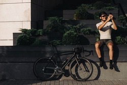 Happy cyclist in sportswear, helmet and glasses using modern smartphone while sitting on city street. Black sport bike standing near. Lifestyles concept.