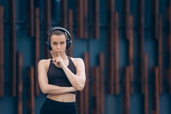 Front view of blonde girl showing quiet. Pretty slim woman standing with crossed arms, listening to music in earphones, looking at camera. Concept of youth and doing sport.