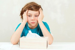 Sick boy sitting at the desk. Kid using paper napkins. Allergic kid, flu season. Box with nose napkins. Boy has a virus, runny nose and headache. Schoolboy blowing runny nose.
