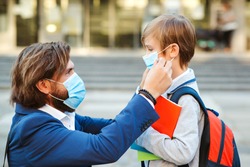 Dad putting on a protective mask on his son's face outdoors. Schoolboy is ready go to school. Cute boy with backpack. Back to school concept. Medical mask to prevent coronavirus. Coronavirus pandemic