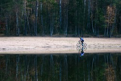 Man on a gravel bike. Cyclist on the empty sandy beach. Autumnal forest and great reflection in water.