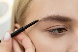 Stylist draws eyebrows with a pencil for a young beautiful woman.