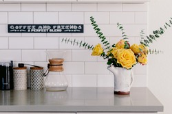 Yellow large roses and eucalyptus branches in the ceramic craft white vase. Kitchen counter top. Lifestyle bright kitchen. Coffee space in the kitchen. Grey and white colors. Bouquet of yellow roses.