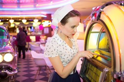 Young woman in retro dress press button of old-fashion musical machine in bar, focus on the machine.