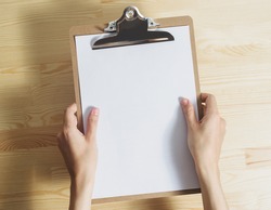top view of white caucasian woman's hands holds clipboard with blank white paper sheet over wooden table