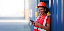 Online working by smartphone. Black female dock worker control loading containers box from cargo at warehouse container yard. Marine and carrier insurance concept. logistic shipping yard