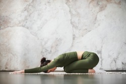 Beautiful young model girl working out at home, doing yoga exercise for spine on black wooden floor, lying in Child Pose, Balasana Posture, resting after practice, side view. Full length, side view
