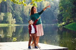 A beautiful mother stands on a lake bridge and points something out to her son with a beautiful landscape of nature.