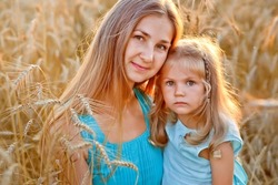 A large portrait of a mother and daughter in blue dresses in a wheat field at sunset. A woman holds a girl in her arms and hugs her against the background of rye ears. They look at the camera.