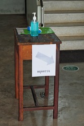 Blue alcohol gel bottle on brown wooden table at the checkpoint before entering the building. The white paper sign has Thai characters meaning 