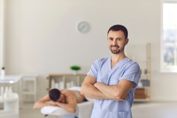 Portrait of happy smiling young professional masseur or manual therapist looking at camera standing arms folded against blurred background of his massage room in modern wellness center