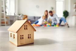 Close-up of miniature detached house on floor at home with happy family playing with little child in background. Mortgage, real estate business, buying property and confidence about future concepts