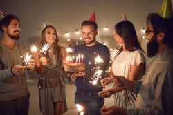 Group of cheerful young people in cone party hats with lit sparklers in hands wishing happy birthday to their friend whos about to blow candles on his birthday cake during celebration at home