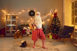 Funny grandpa having fun at Christmas or New Year disco night party at home. Old Santa Claus in polka dot underwear and red and white striped socks holding present and dancing in decorated living-room