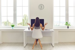 Back view of woman taking break from office work and meditating sitting in modern workspace at table with desktop computer and cup of coffee to go. Concepts of corporate wellbeing and stress relief