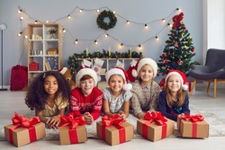 Portrait of a group of happy smiling diverse children lying on the floor near Christmas presents tied with beautiful red bows and looking at camera. Children pose in a room with Christmas decorations.