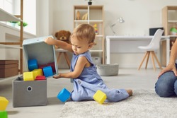 Little helper cleaning up and learning to be independent. Cute 2 year old child putting cubes back in their place after playing. Toddler boy putting toys away sitting on warm floor in nursery room