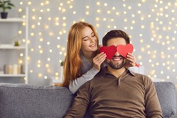 Happy smiling couple in love celebrating their relationship anniversary at home. Young people having fun on Saint Valentine's Day. Woman covering boyfriend's eyes with two red heart-shaped cards