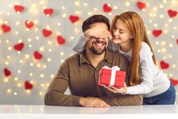 Young couple in love celebrating Saint Valentine's Day or relationship anniversary. Happy woman covering boyfriend's eyes giving him surprise gift. Smiling man getting present from loving girlfriend