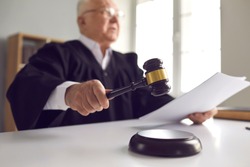 Stern judge with paper document pronouncing sentence in a court of law. Judge finds the accused guilty, passes judgement and rules case closed. Hand holding gavel and hitting sound block in close-up