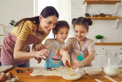 Happy family busy in kitchen. Mother and two helpful little daughters in aprons making cookies on weekend at home. Mom teaches inquisitive children to break egg, prepare dough and use cooking utensils