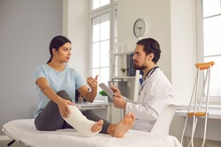 Physical injury treatment: Serious doctor listening to patient with bone fracture. Young woman with broken leg talking to traumatologist or orthopedic surgeon during medical exam in hospital or clinic