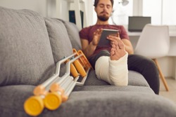 Young man with broken leg in plaster cast sitting on sofa and using tablet computer. Close up of foot and crutches. Physical injury, bone fracture, treatment and rehabilitation at home concept