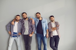 Male friendship, company, friends concept. Group of four smiling positive young men friends in casual clothes standing hugging and looking at camera over grey wall background at home