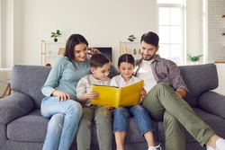 Happy young family with children enjoying free time resting on couch at home. Parents and kids sitting on soft comfy sofa, reading book of fairy tales or looking through holiday photo album together