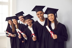 Smiling international students in traditional academic caps and gowns celebrating graduation. Group portrait of happy multiethnic college or university graduates holding diplomas and looking at camera