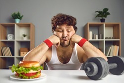 Upset depressed young male athlete looking at dumbbell and tempting burger choosing either sport or fast food. Weight loss, fitness, will power, choice between healthy and unhealthy lifestyle concept