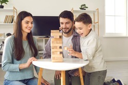 Happy family with child taking blocks from tumble tower. Smiling young Caucasian couple with little son enjoying free time on weekend at home, having fun and playing exciting board game together