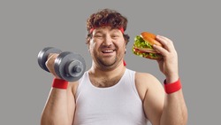 Funny smiling man with dumbbell and delicious burger looking at camera. Happy fat guy holding free weights and eating big yummy hamburger. Sport, food, failed diet, workout exercise, cheat day concept