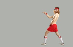Funny weird freak foolish topless shirtless bare chested thin skinny young man in glasses, red gym headband and sports shorts standing on grey background, pointing with finger to advertising copyspace