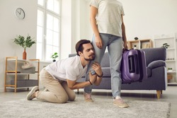 Young married couple breaking up. Angry woman leaving home with packed suitcase. Clingy desperate husband on floor holding wife's leg begging his love to stay. Relationship breakup and divorce concept
