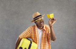 Studio portrait of happy man ready for flight. Black guy in glasses, orange shirt and summer hat holding yellow travel suitcase and paper airplane smiling at camera isolated on gray background
