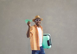 Happy tourist ready for flight. Black guy in glasses and summer hat walking to board plane. Portrait of man with travel suitcase and paper airplane in hands smiling at camera on gray studio background