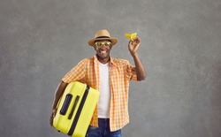 Portrait of happy man ready for flight. Smiling black guy in glasses, orange shirt and summer hat holding yellow travel suitcase and paper airplane looking at camera standing on gray studio background