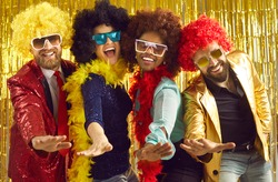 Pop music band singing at night club. Young people disguised in fancy feather boas and funny silly curly wigs dancing together. Happy multiethnic men and women having fun at Halloween disco party