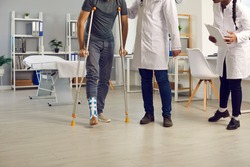 Appointment with orthopedic surgeon. Patient with broken leg or ankle sprain stands up from examination bed and walks on crutches assisted by diverse hospital staff of professional doctors and nurses