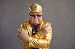 Retired older adult man in thug life glasses, gold chain and disco outfit isolated on gray background. Studio portrait senior pensioner in funny sunglasses looking at camera with angry face expression