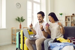 Happy couple getting ready for tour vacation trip pay for cheap hotel online. Excited boyfriend and girlfriend sitting on sofa with laptop, credit card, passports, flight tickets and travel suitcase