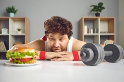 Funny upset depressed fat athlete choosing between gym dumbbell and delicious cheeseburger. Concept of sport, choice, willpower, healthy weight loss fitness exercise vs yummy unhealthy fast food diet