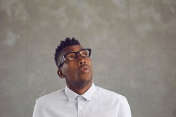 Wow that's interesting. African man sees something curious. Young black guy in white shirt and stylish eyeglasses looks up and whistles surprised by unexpected news on gray copy space background above