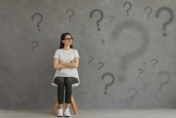 Serious confused woman in glasses sitting on chair looking at multiple question marks on grey background. University, college, high school teen girl doubting, searching, choosing interesting solution
